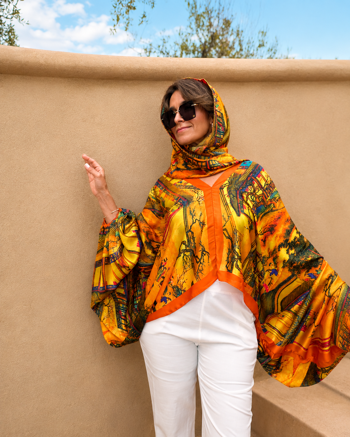 A woman wearing a vibrant yellow and orange patterned headscarf and matching long-sleeve shawl top with white pants, posing against a beige stucco wall under a blue sky.