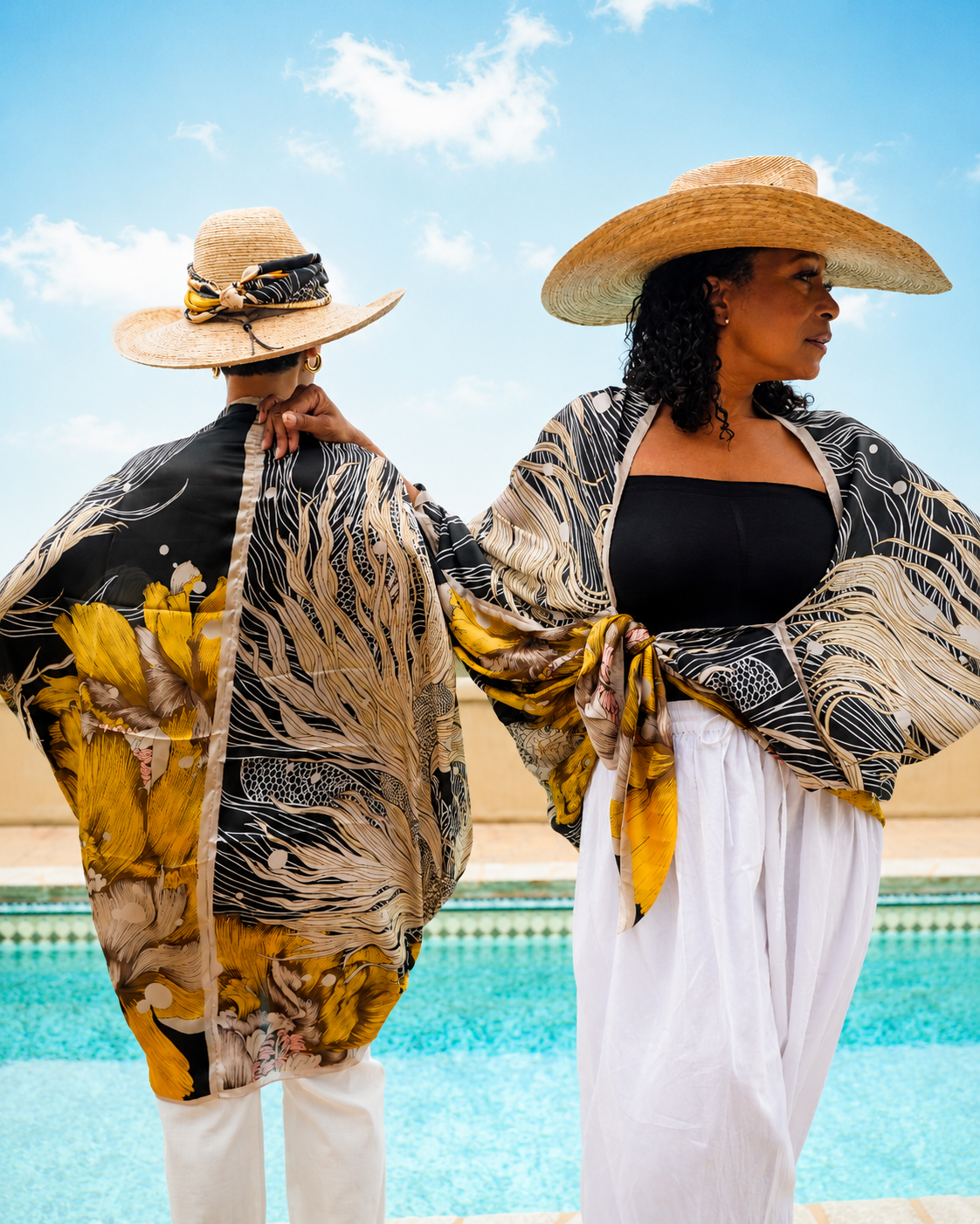 Two women standing by a swimming pool wearing matching wide-brim straw hats and a matching black, beige, and mustard yellow abstract-patterned kimono-style cover-up shawl. One woman is shown from the back and the other from the side, both wearing white pants.