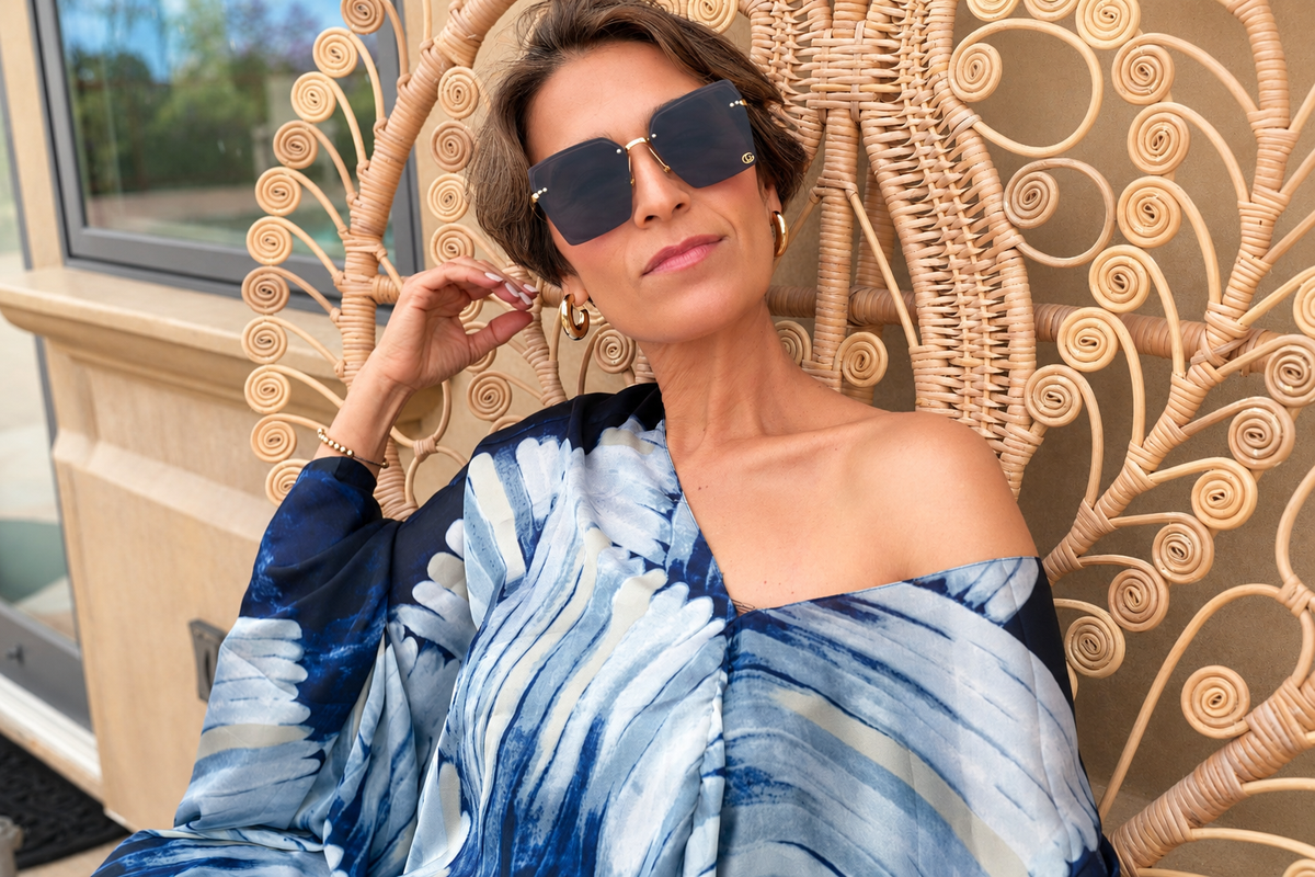A woman wearing oversized dark sunglasses and a blue, white, and gray patterned off-the-shoulder shawl top, leaning against a decorative rattan chair.